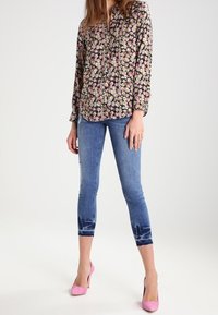 Blusa con stampa floreale a maniche lunghe, sfondo nero, fiori rosa e gialli; jeans capri in denim blu con dettagli strappati; tacchi rosa.