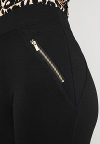 Mustat legginsit, joissa on sivutasku kullalla varustetulla vetoketjulla. Ne on valmistettu sileästä, joustavasta kankaasta ja niissä on istuva muoto.