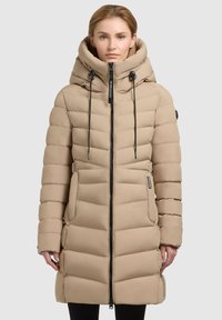 Beige pufferjas met een hoge kraag, ritssluiting en trekkoorden. Beschikt over een gestikt ontwerp en zijzakken. Glad textiel.