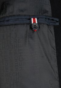 Tejido negro con letras en relieve, que cuenta con un botón y un acento a rayas rojas y blancas. La textura es suave con costuras visibles.