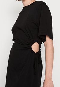 Abito nero con maniche corte e larghe, caratterizzato da un lato arricciato con un'apertura ovale e un dettaglio con cordoncino in vita. Tessuto dalla texture liscia.