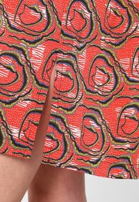 Punainen kangas, jossa on toistuva abstraktin kuvio, johon kuuluu mustalla ja keltaisella viivatut pyöreät muodot. Taustalla on myös valkoisia raitoja ja teksturoituja yksityiskohtia.