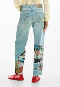 Blugi din denim albastru deschis cu o scenă costieră pictată pe picioare, reprezentând clădiri și un pod. Include buzunare decorative în spate.