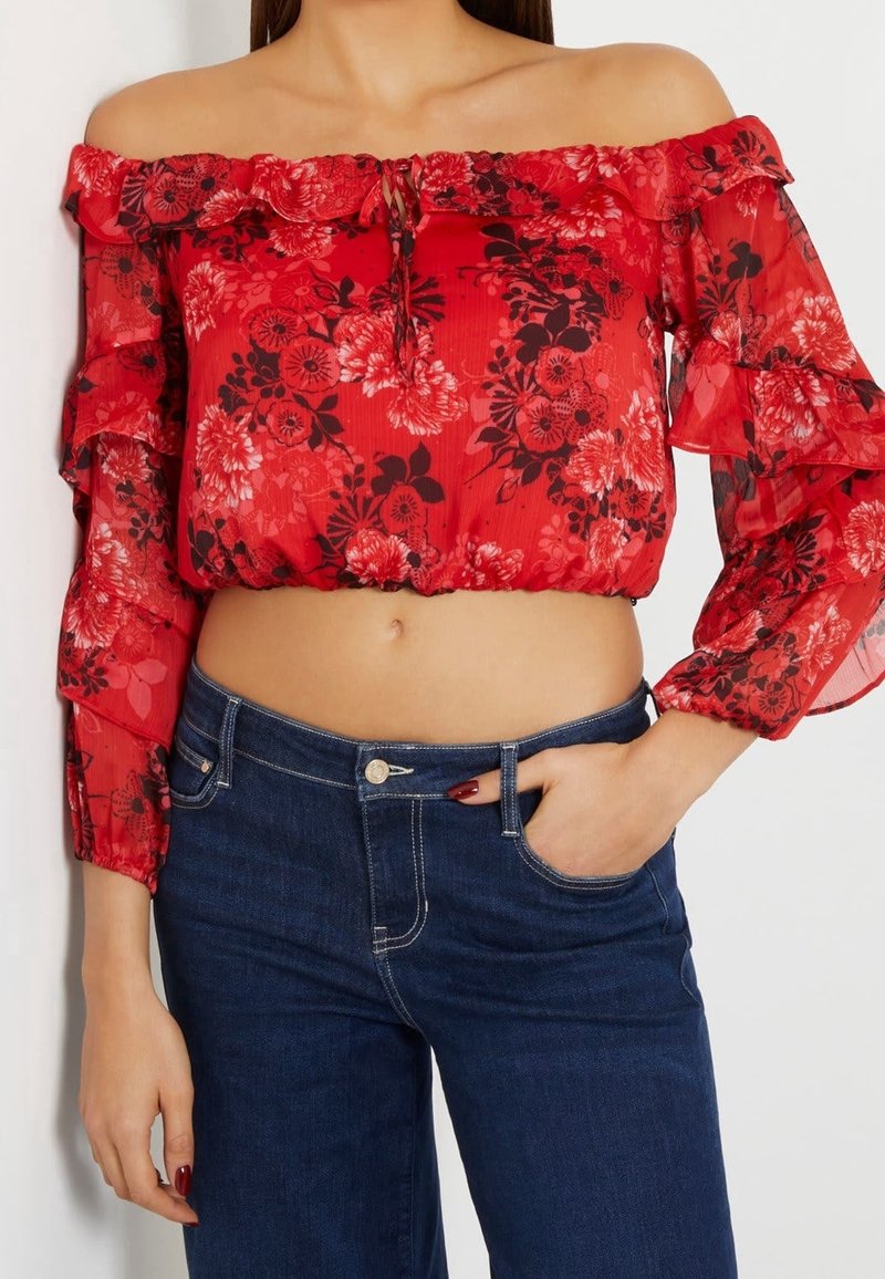 Femme portant un crop top rouge à fleurs à épaules dénudées avec manches volantées et un jean bleu foncé, main dans la poche sur fond blanc.