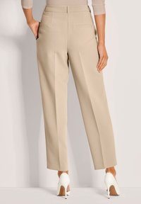 Vrouw in beige op maat gemaakte rechte broek en witte hoge hakken, van achteren gezien met de handen langs de zij.