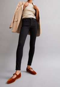 Svarta smala jeans i kombination med en beige blazer och en krämfärgad topp, kompletterade med orange sammetsskor med rem och spänne.