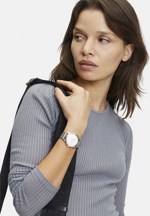 Silberne Armbanduhr mit einem runden Ziffernblatt und einem metallischen Armband, kombiniert mit einem grauen, gerippten Langarmoberteil, gehalten von einem schwarzen Riemen.