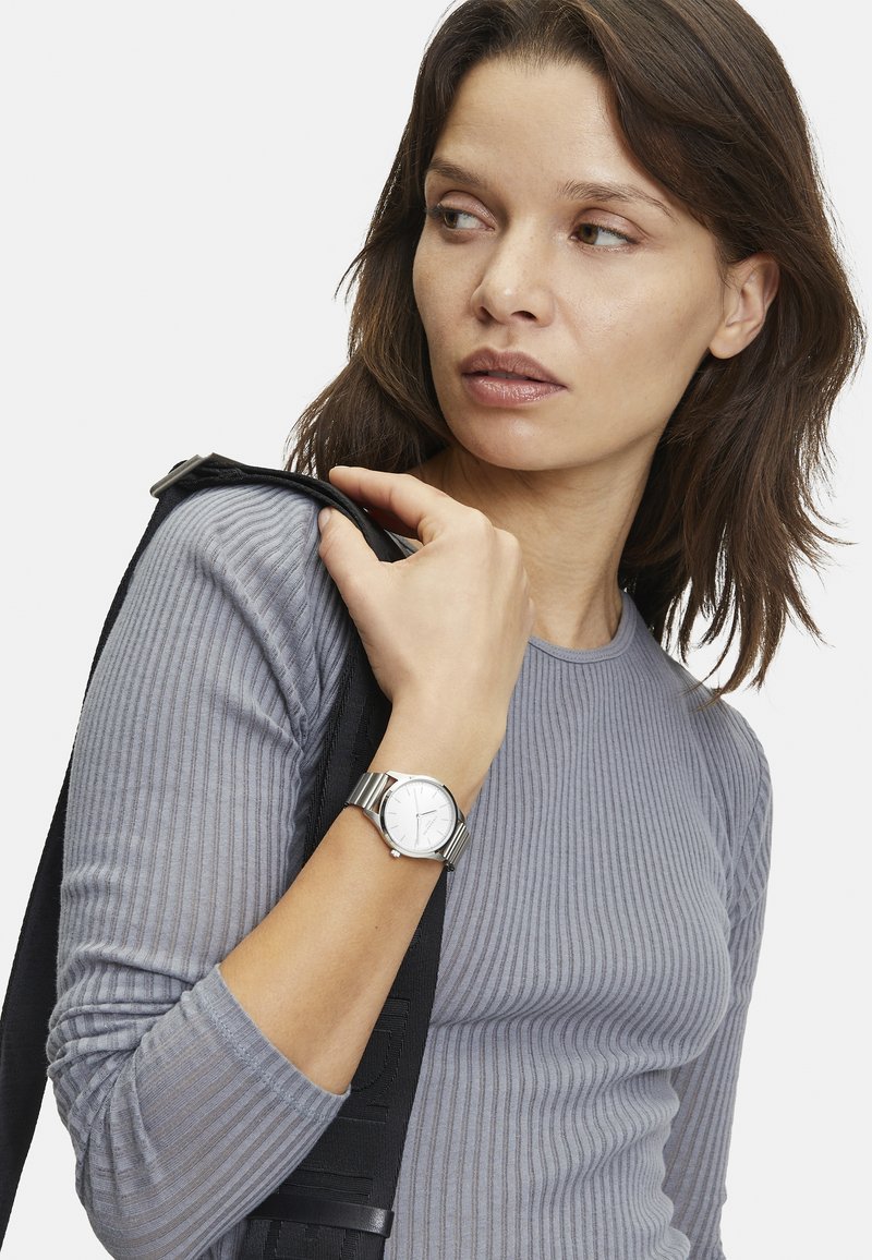 Silverfärgad armbandsur med rund urtavla och metallarmband, tillsammans med en grå, ribbad långärmad topp, som hålls av ett svart band.