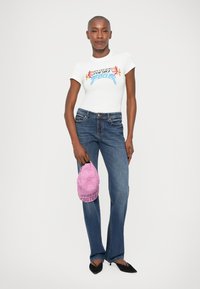 Biely tričko s farebným grafickým dizajnom, tmavomodré džínsy a čierne špicaté baleríny. Drží ružovú čiapku s potlačou "Versace Jeans Couture".