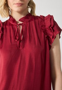 Rød bluse med volanger p�å korte ermer, nøkkelhull-utsnitt med knytedetalj, laget av glatt stoff, som gir en flytende fall og teksturert finish.