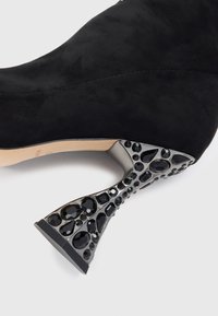 Cizmă cu toc înalt din suede negru, având un toc stiletto sculptat și lucios de culoare neagră, cu un aranjament decorativ de bijuterii negre și accente texturate.