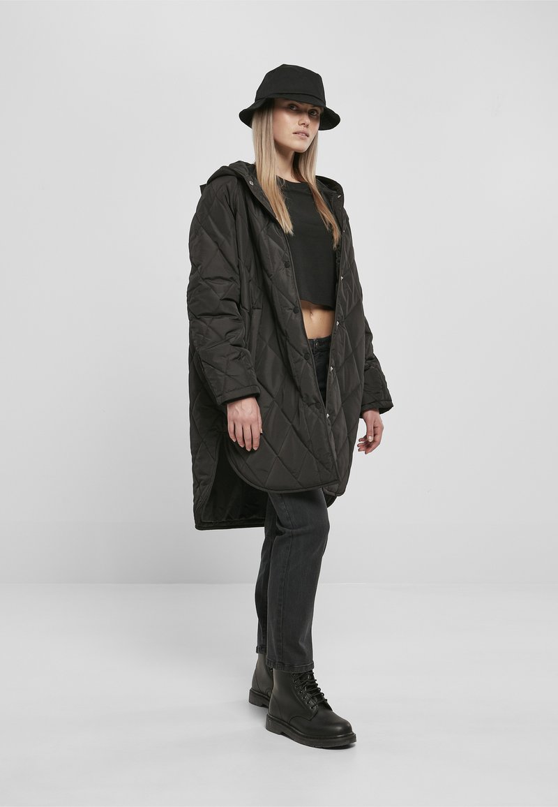 Žena nosi čierny prešívaný kabát, čierny bucket hat, krátky čierny top, tmavé džínsy a čierne bojové topánky, stojí pred sivým pozadím.