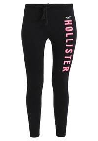 Czarne spodnie dresowe slim-fit z różowym pionowym napisem "HOLLISTER" oraz małym białym logo mewy na prawym udzie, z elastycznym paskiem w pasie z sznurkiem.