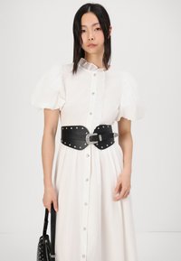 Vit skjortklänning med korta puffärmar, rynkad krage och svart läderbälte med nitar. Modell som håller en svart handväska.