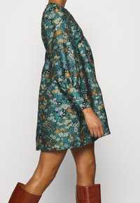 Blumengemustertes Kleid aus türkisfarbenem Stoff mit Puffärmel, Rundhalsausschnitt und ausgestellter Silhouette, kombiniert mit kniehohen braunen Stiefeln.