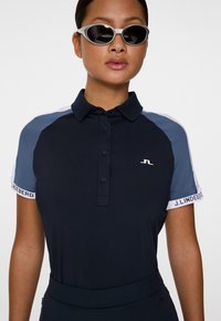 Granatowa koszulka polo z kontrastującymi jasnoniebieskimi i białymi rękawami, kołnierzykiem na trzy guziki oraz haftowanym logo na piersi. Noszona z białymi okularami przeciwsłonecznymi.