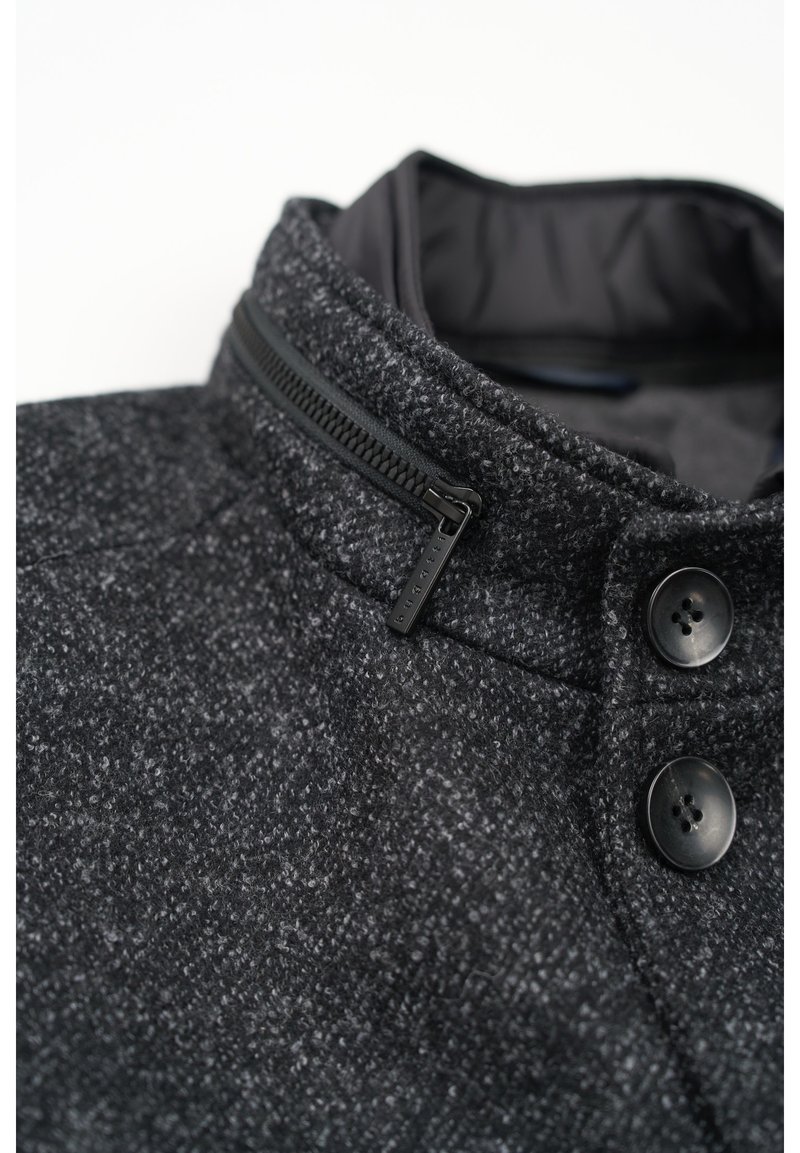 Giacca grigia con texture e colletto alto, dotata di una zip nera e due bottoni neri. Fodera interna liscia visibile al colletto.