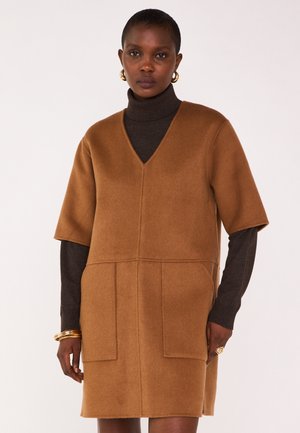 Vestito marrone in misto lana con maniche corte, scollo a V profondo e due tasche frontali. Sotto si intravede un dolcevita scuro. Dettaglio di bracciale dorato.