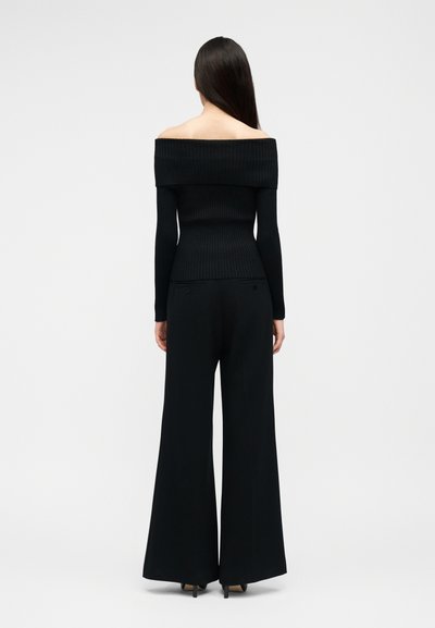 Vrouw met lang donker haar, gekleed in een zwart off-shoulder geribd topje en zwarte wijde broek, staand met de rug naar de camera tegen een effen achtergrond.