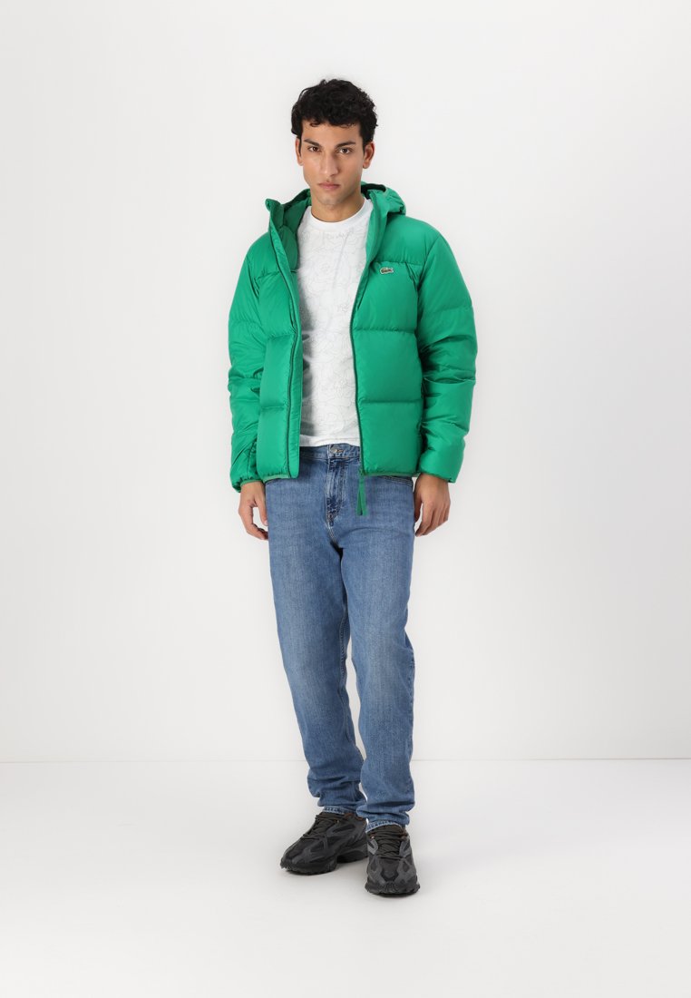 Veste à enfiler verte avec un design matelassé, fermeture éclair et capuche, associée à un jean bleu clair et des baskets noires sur un fond uni.