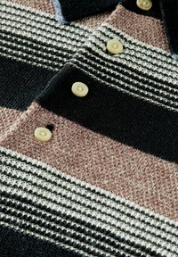 Gros plan d’un vêtement tricoté avec des rayures horizontales noires, beiges et blanches et quatre boutons beiges au centre.