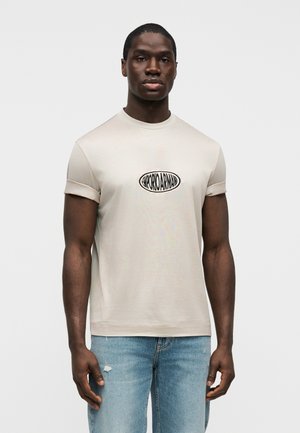 Mann trägt ein beigefarbenes kurzärmeliges T-Shirt mit hochgekrempelten Ärmeln und einem schwarzen ovalen Logo auf der Brust, kombiniert mit hellblauen, abgenutzten Jeans.