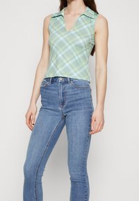 Zielona w kratkę top bez rękawów z kołnierzykiem, z dekoltem w kształcie litery V. Połączony z dopasowanymi niebieskimi dżinsami o klasycznej teksturze denimu.