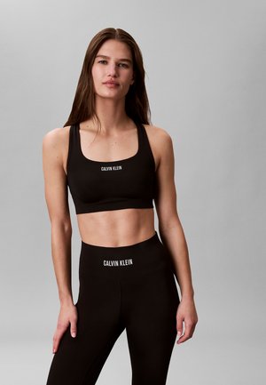 Kobieta w czarnym sportowym biustonoszu Calvin Klein i legginsach z wysokim stanem, stojąca na jasnoszarym tle.