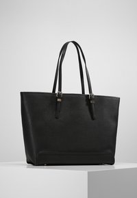 Černá kožená taška přes rameno s dvojitými ramenními popruhy a stříbrnými sponami, vystavená na bílém podstavci na neutrálním pozadí.