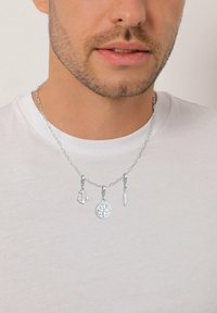 Man med silverhalsband smyckat med en ankare, kompass och fjäderberlocker över en vit rundhalsad tröja.