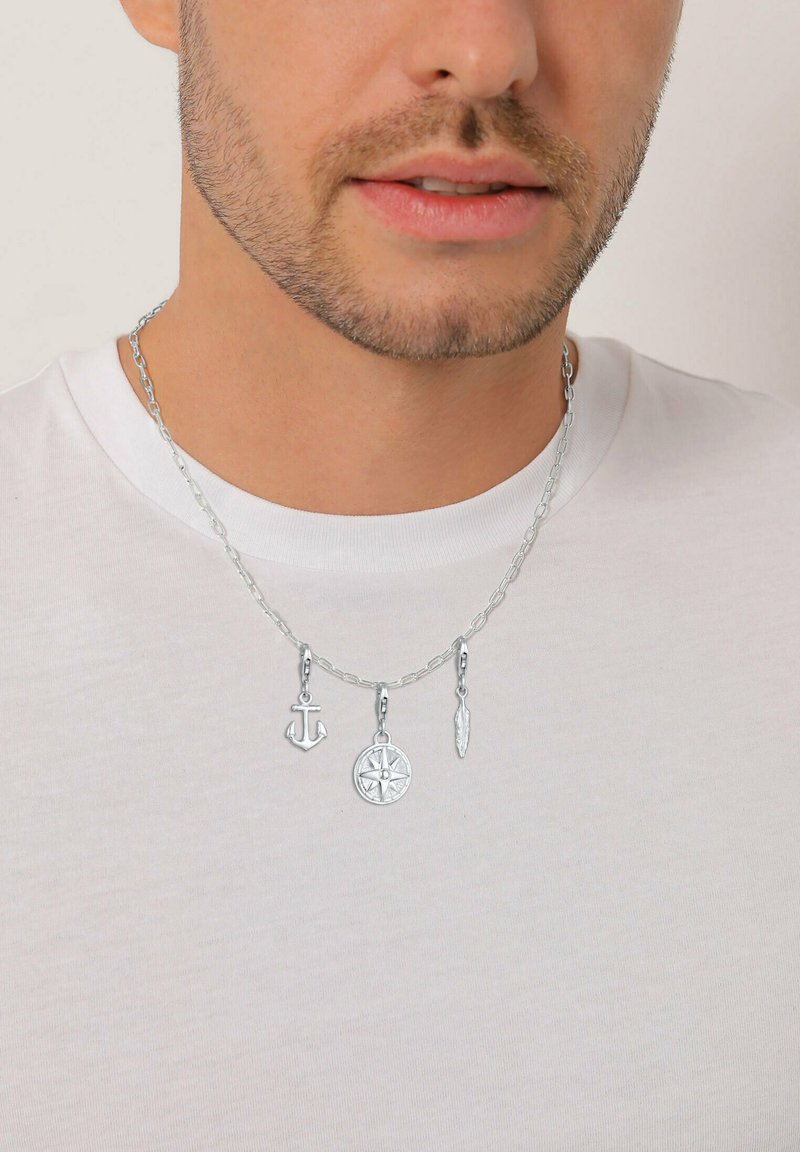 Man med silverhalsband smyckat med en ankare, kompass och fjäderberlocker över en vit rundhalsad tröja.