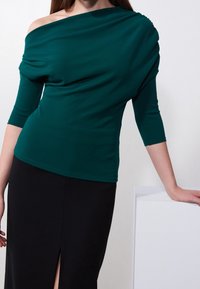 Teal, draperad off-shoulder topp med tre fjärdedelars ärmar i slät tyg, kombinerad med en svart kjol med hög slits fram.