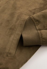 Manche de veste en daim marron avec bord-côte tricoté et poche latérale inclinée visible sur un fond uni.