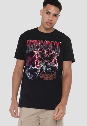 Schwarzes Baumwoll-T-Shirt mit einem grafischen Aufdruck eines Demogorgons aus "Stranger Things" mit roten und violetten Akzenten und fettem Text.