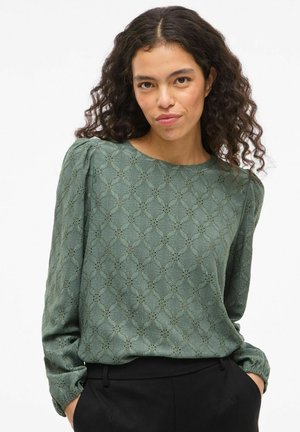 Zaļa garām piedurknēm blūze ar teksturētu rakstu, ar pūkainām plecu daļām un apaļu kakla izgriezumu, apvienota ar melnajām biksēm.