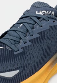 Running shoe with a textured, dark blue synthetic upper, featuring gray laces and a bold yellow sole. Includes branding on the tongue.