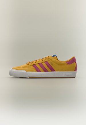 Žltá teniska Adidas s ružovými pruhmi, bielou podrážkou a šnúrkami, zobrazená z boku na neutrálom pozadí.