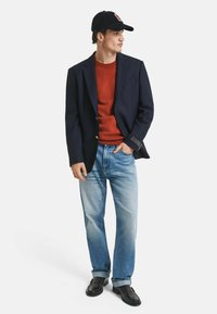 Marineblazer, roter Pullover, hellblaue Jeans, schwarze Schuhe. Der Blazer hat goldene Knöpfe; der Pullover hat eine glatte Textur; die Jeans sind leicht ausgebleicht.