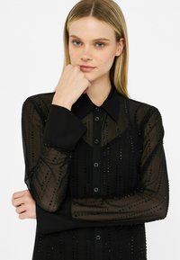 Blouse noire transparente avec des manches longues, fermeture à boutons à l'avant et col structuré. Présente des motifs de perles verticales pour la texture et le détail.
