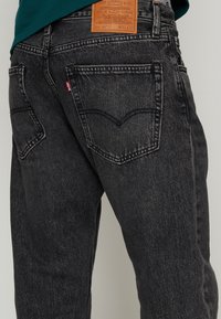 Jeans de mezclilla negra con corte ajustado, que cuentan con una etiqueta de cuero, bolsillos traseros curvados y detalles de costura sutiles. La tela tiene una ligera textura.
