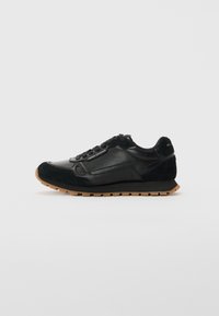 Baskets en cuir noir et en daim avec semelle en caoutchouc clair et logo "Armani Exchange" sur le côté et le talon, présentées de profil sur fond blanc.