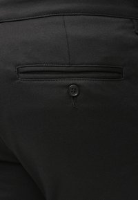 Schwarze Hose mit einer Gesäßtasche, die mit einem einfachen Knopfverschluss und dezenten Steppnähten versehen ist. Das Material wirkt strukturiert und strapazierfähig.