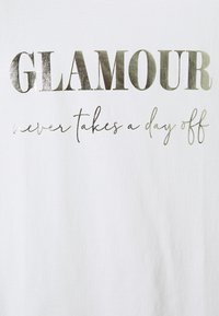 Camiseta blanca de manga corta con un atrevido texto metálico plateado: "GLAMOUR" en mayúsculas y "nunca se toma un día libre" en estilo cursivo.