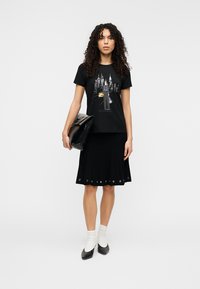 Μαύρο βαμβακερό μπλουζάκι με γραφικό τύπωμα που περιλαμβάνει αστικά στοιχεία. Συνδυάζεται με μαύρη φούστα με πιέτες και μαύρα τακούνια. Αξεσουάρ περιλαμβάνουν μια μαύρη τσάντα χειρός.