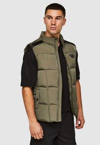 Olivengroen puffervest met een ritssluiting aan de voorkant, horizontaal gestikte design en zwarte schouderaccenten, gedragen over een zwart T-shirt.