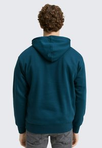 Teal hoodie met ribbels aan de manchet en onderrand, met een zachte textuur. De achterkant toont de capuchon en een gladde stofafwerking.