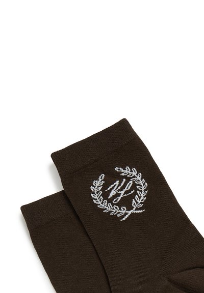 Chaussettes en tricot marron avec couronne de laurier blanche brodée et initiales stylisées sur la zone de la cheville, posées à plat sur fond blanc.