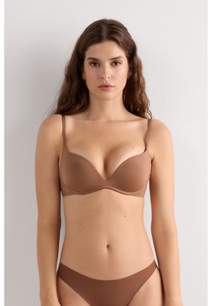 Femme aux longs cheveux ondulés portant un soutien-gorge et une culotte marron assortis, debout contre un mur blanc uni, regardant devant elle.