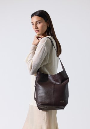 Mujer con suéter claro y falda lleva un gran bolso de cuero marrón oscuro al hombro, mirando por encima del hombro contra un fondo liso.