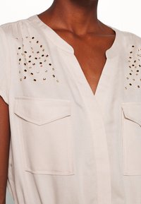 Blouse beige avec col en V, manches courtes et deux poches avant. Dotée de clous métalliques décorant les épaules selon un motif dégradé.